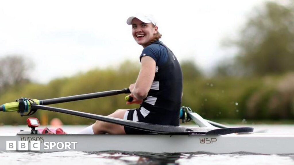 Lauren Henry turns and smiles while in her boat on the water, wearing a dark rowing outfit and white cap