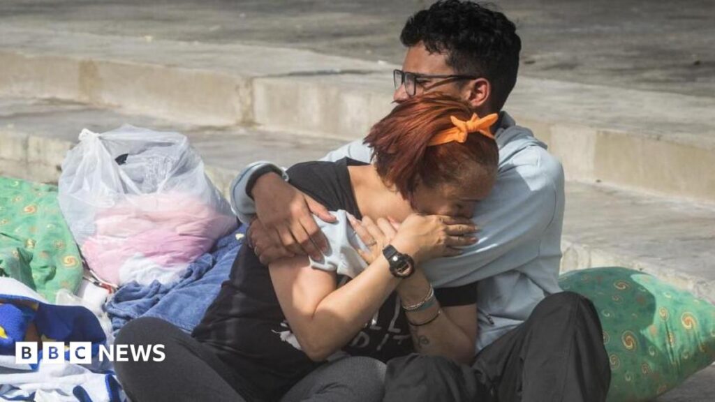 Two people cry into each others' arms surrounded by pillows, blankets and carrier bags of possessions on concrete stairs outside