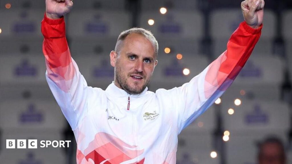 Sam Tolchard, wearing a white and red England tracksuit top, raises his arms while standing on the podium at the 2026 Commonwealth Games in Glasgow