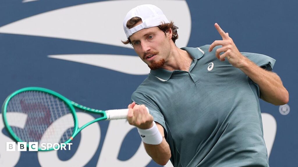 Toby Samuel, wearing a grey shirt and white backwards baseball cap, hits the ball with a green racquet