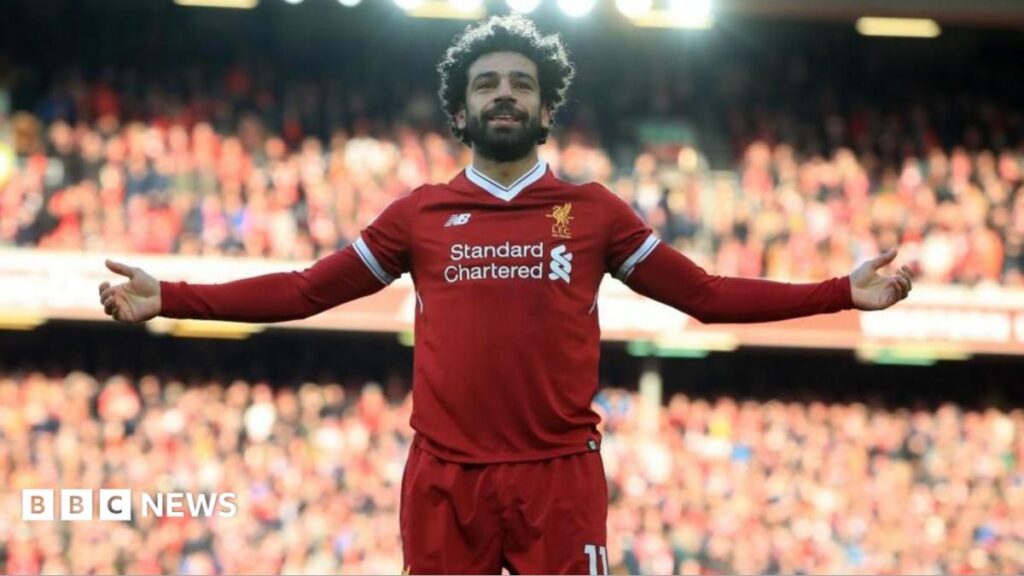 A man with black hair and a beard is wearing a Liverpool kit while standing on the pitch with his arms raised to either side. He is smiling. A large crowd can be seen behind him.