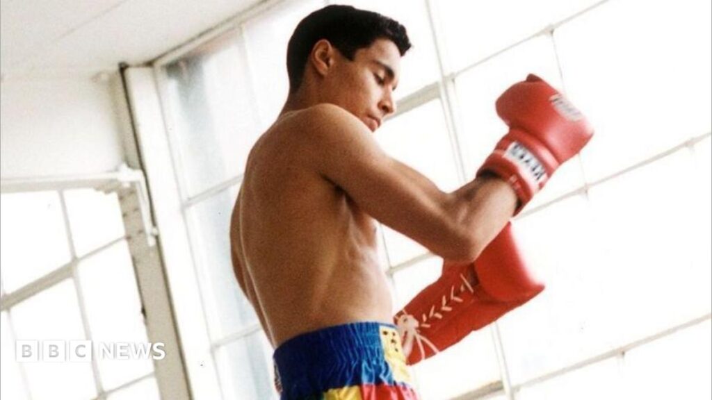 A young boy shadow boxed in what looks like an empty warehouse with bright natural light shining into the room. He is wearing blue, yellow and red shorts and red gloves.