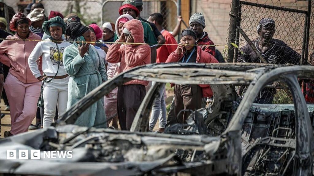 A crowd of people look on close to a burned out car