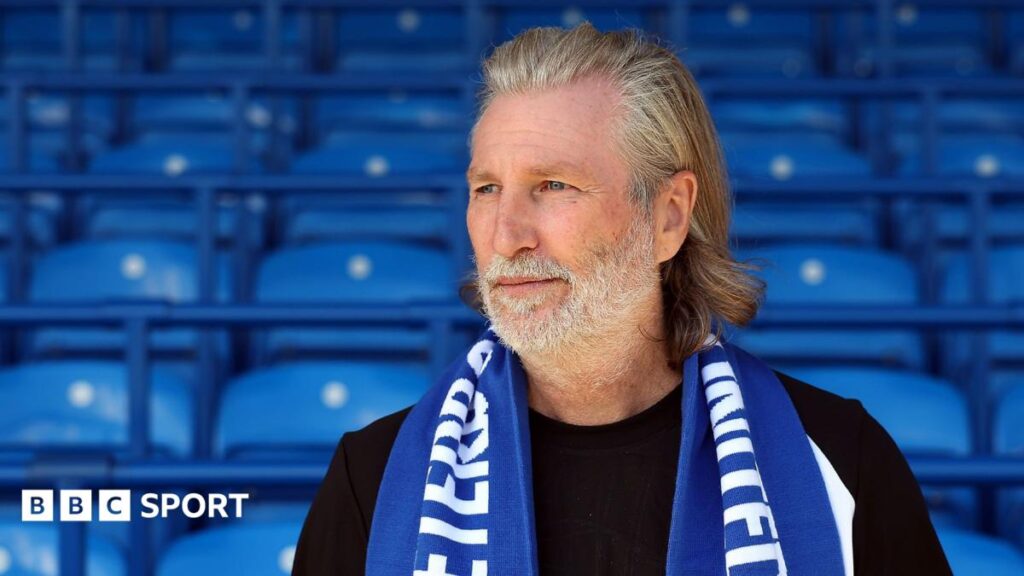 Robbie Savage in a black T-shirt poses with a blue Peterborough scarf